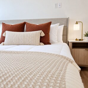 A neatly made bed with a white comforter and multiple pillows, a bedside table with a lamp, and a wooden nightstand. - Modern Downtown SLC | King Bed + Free Parking (Salt Lake City)