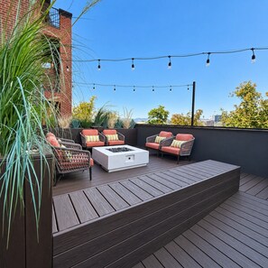 Eine Terrassenausstattung im Freien mit Korbmöbeln, einem weißen Feuerkorb und Lichterketten vor blauem Himmel.