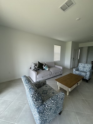 A living room with a grey sofa, patterned armchairs, a wooden coffee table, and a window with blinds. - New 3BR Townhome in Naples - Resort Pools & Gym (Naples)