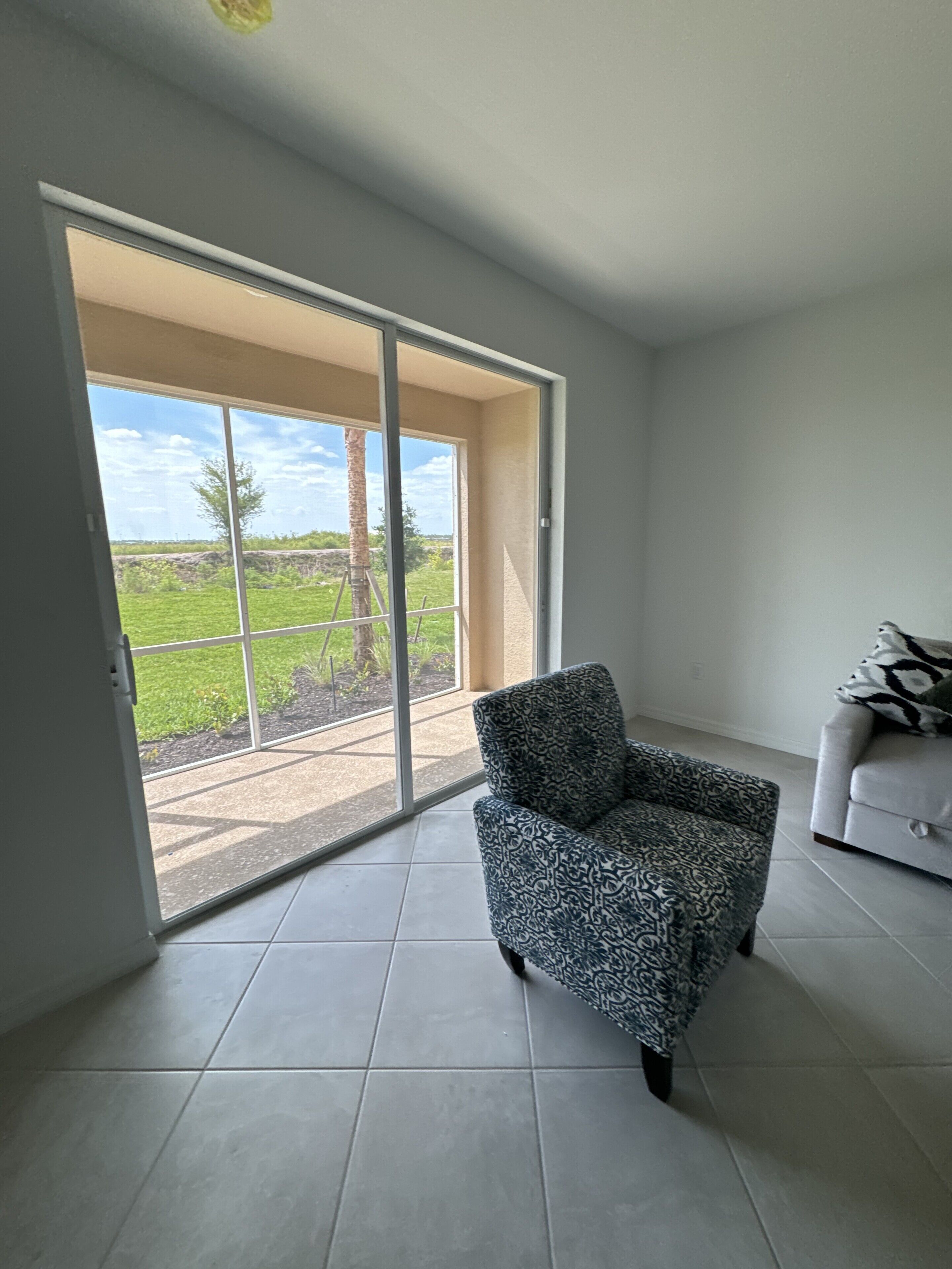 A room with a patterned armchair, a sofa, and a sliding glass door leading to a patio with a view of greenery and a clear sky.