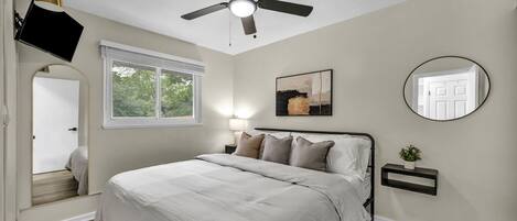 Un dormitorio con cama, ventilador de techo, ventana, televisor montado en la pared, mesita de noche con lámpara y espejo.