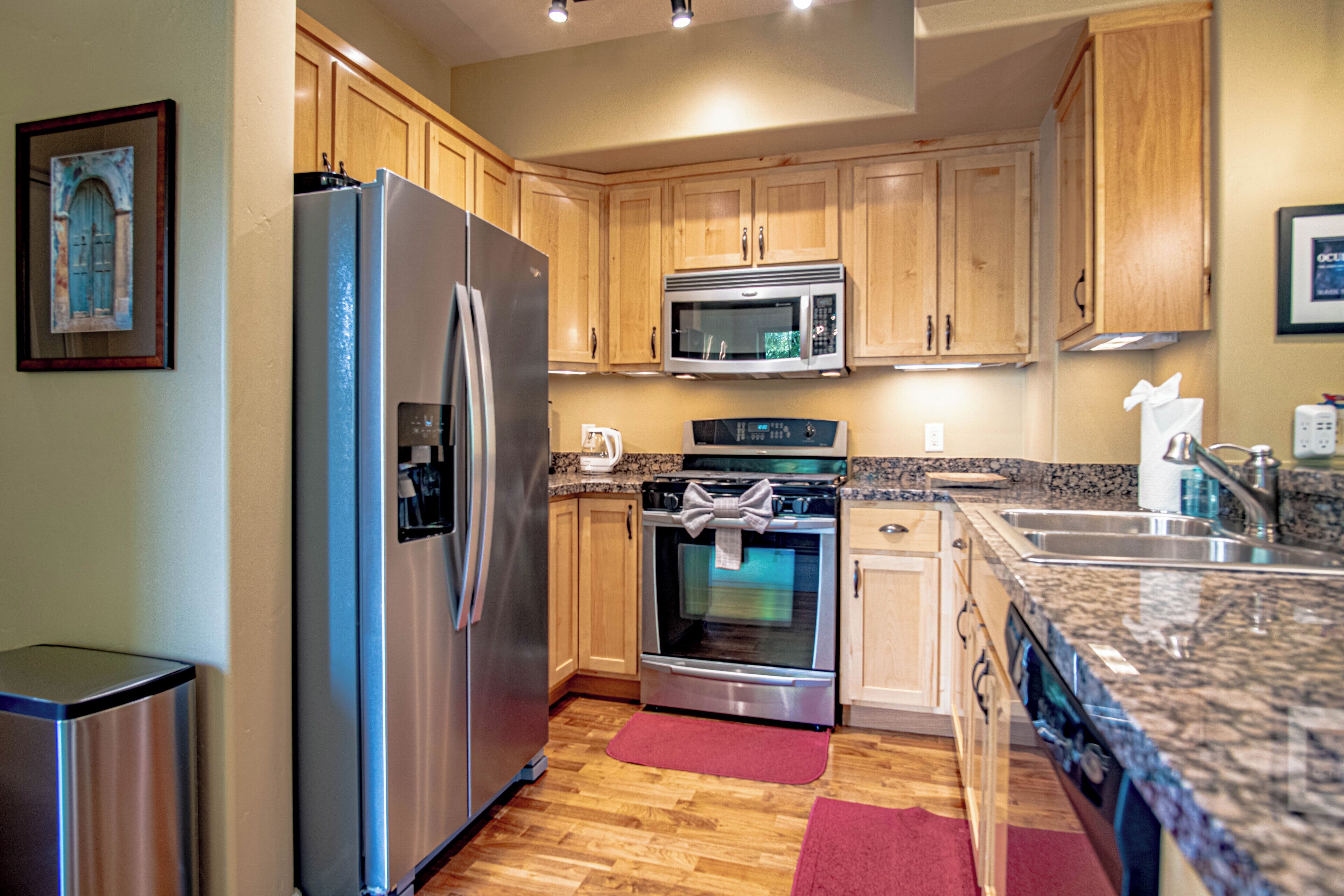 A modern kitchen with stainless steel appliances, wooden cabinets, granite countertops, and a red rug.
