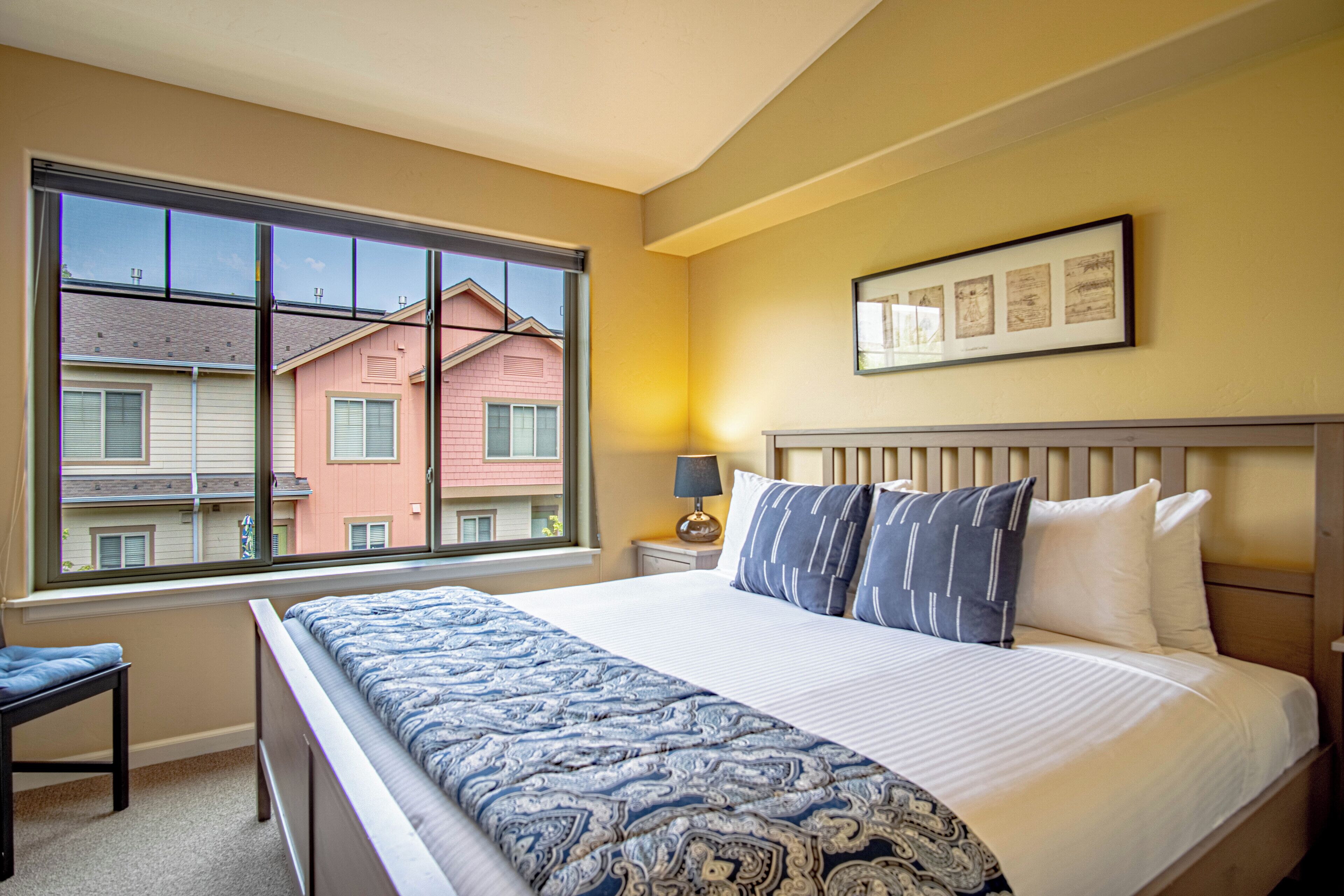 A hotel room with a large bed, a nightstand, a lamp, a chair, and a window with a view of neighboring buildings.