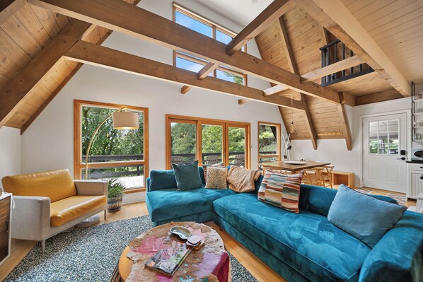Ein großzügiger Wohnbereich mit hohen Decken und Holzbalken, einem blauen Sofa, einem Holztisch und großen Fenstern mit Blick auf Grünflächen.