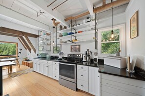 A modern kitchen with white cabinetry, a black countertop, and a built-in oven. There are shelves above the counter holding various kitchen items, and a window with a view of greenery outside. - Private Mountain Getaway Near Boone with Hot Tub (Vilas)