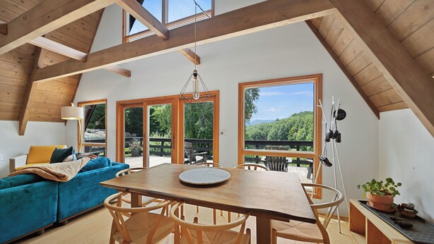 Ein großzügiges Loft mit hohen Decken, Holzbalken, einem Essbereich mit Tisch und Stühlen, einem blauen Sofa und großen Fenstern mit Blick auf Grünflächen.