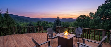 A deck with a fire pit table and chairs, surrounded by a wooden deck and a view of the mountains.