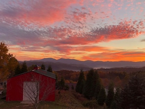 Ein rotes Bauernhaus mit weißer Tür, umgeben von Bäumen und Bergen im Hintergrund.