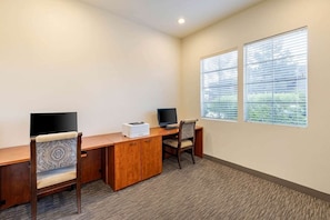 A room with a wooden desk, two chairs, a printer, and a television. There are two windows with blinds.