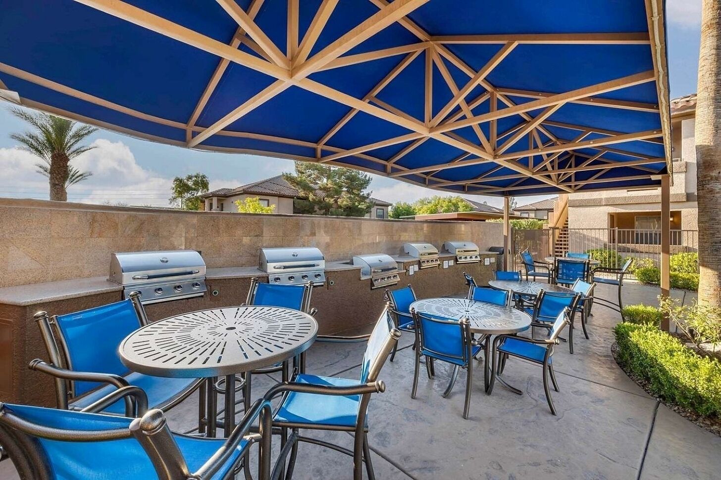 An outdoor patio with round tables and blue chairs, a barbecue grill, and a covered area with a blue roof.