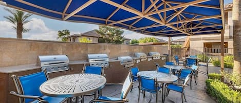 An outdoor patio with round tables and blue chairs, a barbecue grill, and a covered area with a blue roof.