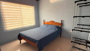 Une chambre avec un plafond en bois, un lit recouvert d’une courtepointe bleue, une fenêtre avec des stores et une étagère en métal.