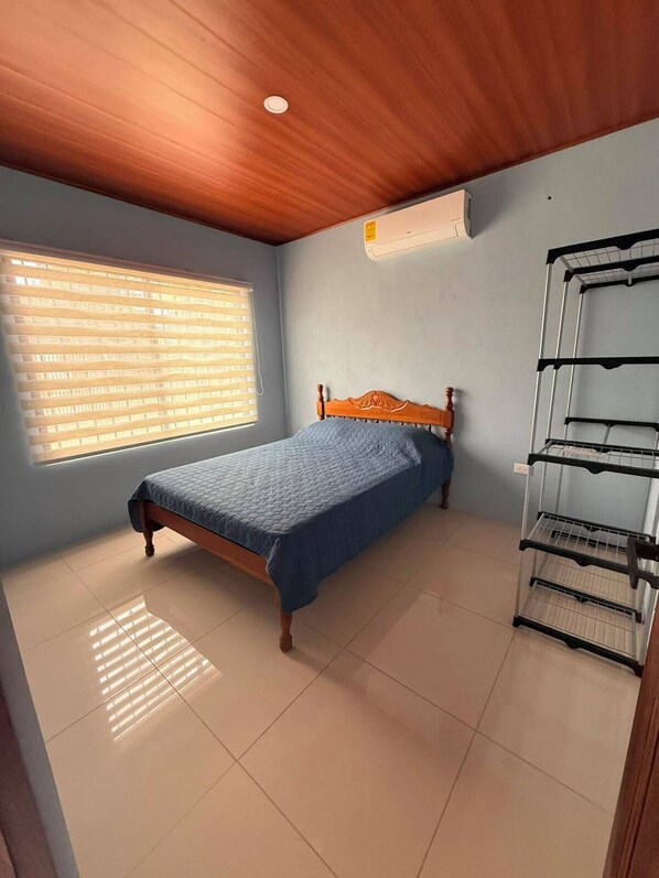 A bedroom with a wooden ceiling, a bed with a blue bedspread, a window with blinds, and a metal shelf unit.