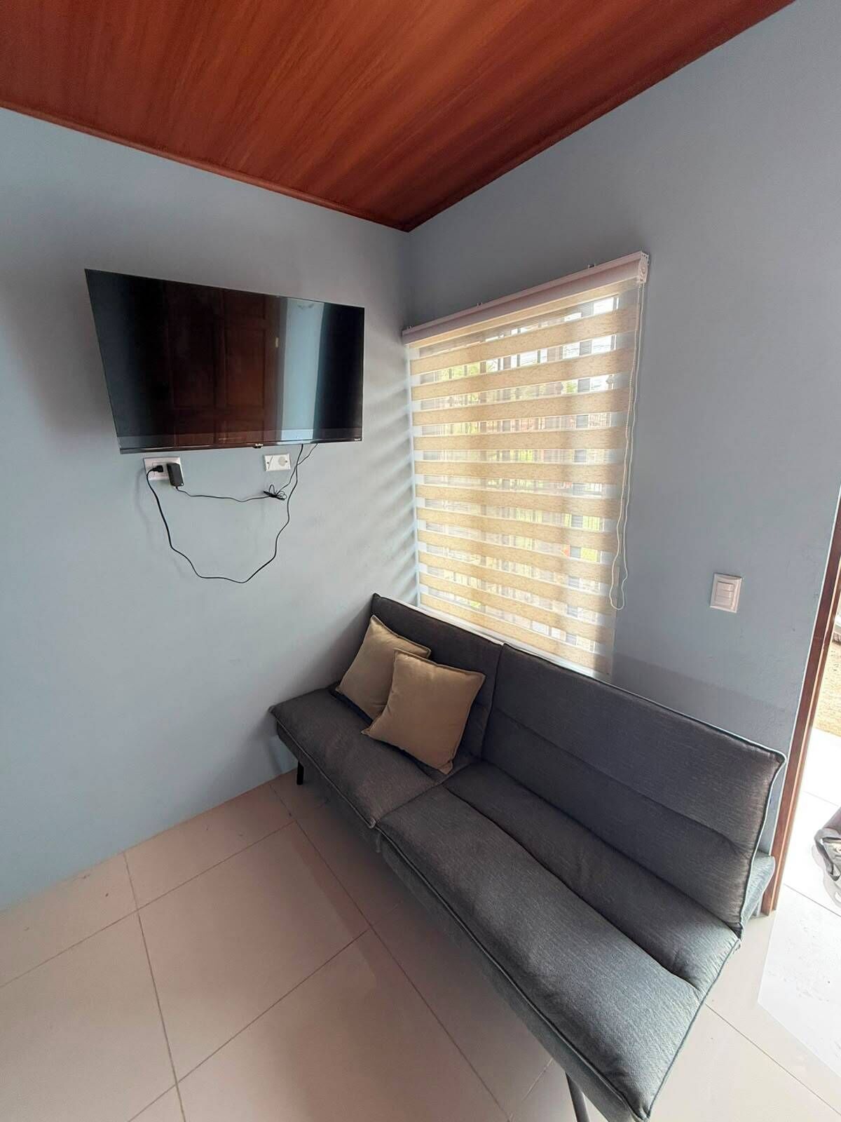 A room with a grey sofa, a mounted flat-screen TV, and a window with blinds.