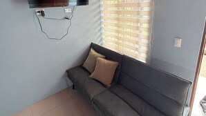 Une pièce avec un canapé gris, un téléviseur à écran plat fixé au mur et une fenêtre avec des stores.