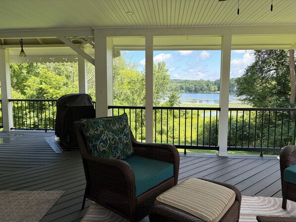 A covered outdoor deck with wicker furniture, a grill, and a view of a lake.