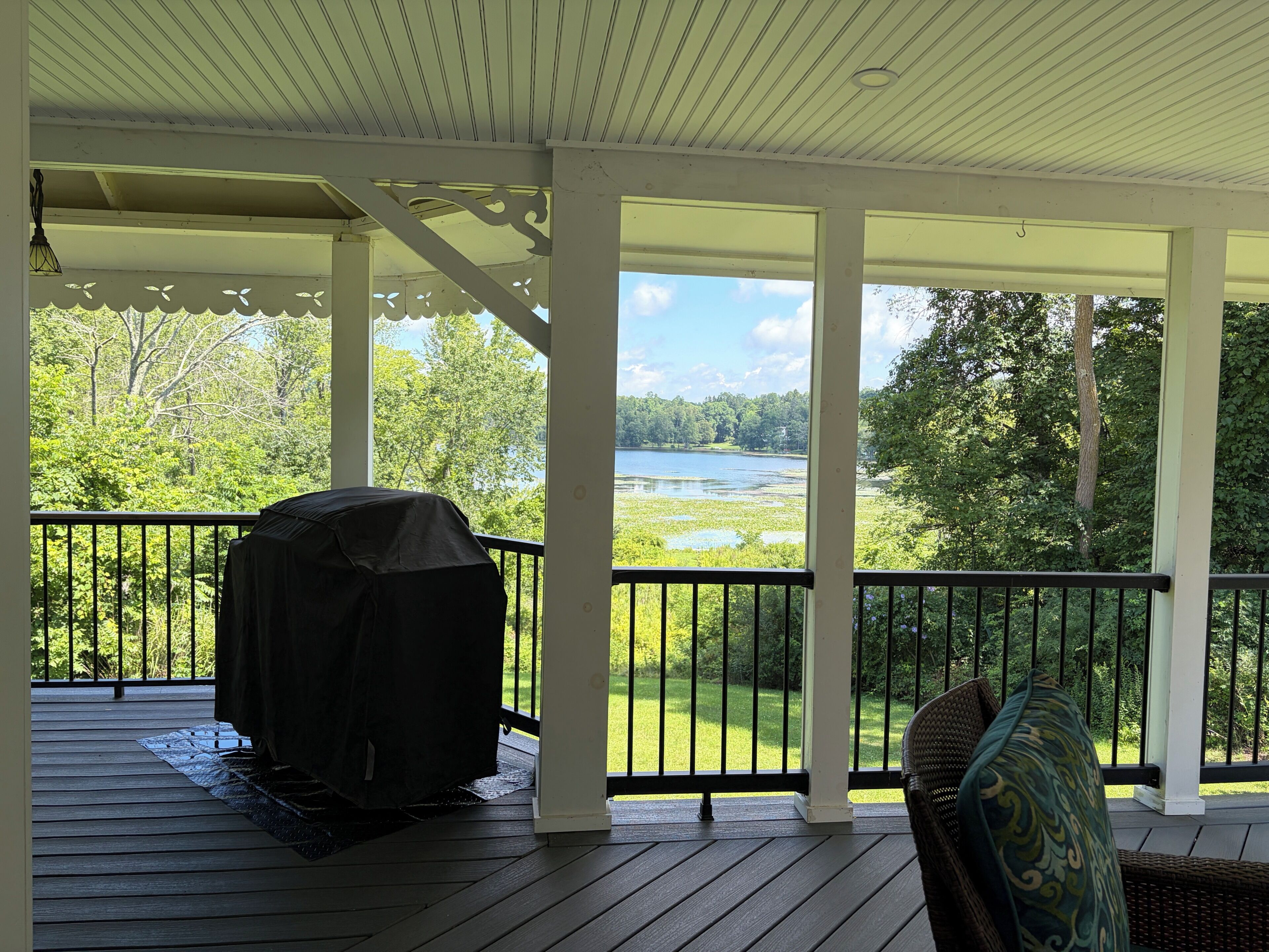 A covered outdoor deck with a grill, a chair, and a view of a lake and forest.