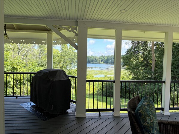 A covered outdoor deck with a grill, a chair, and a view of a lake and forest.