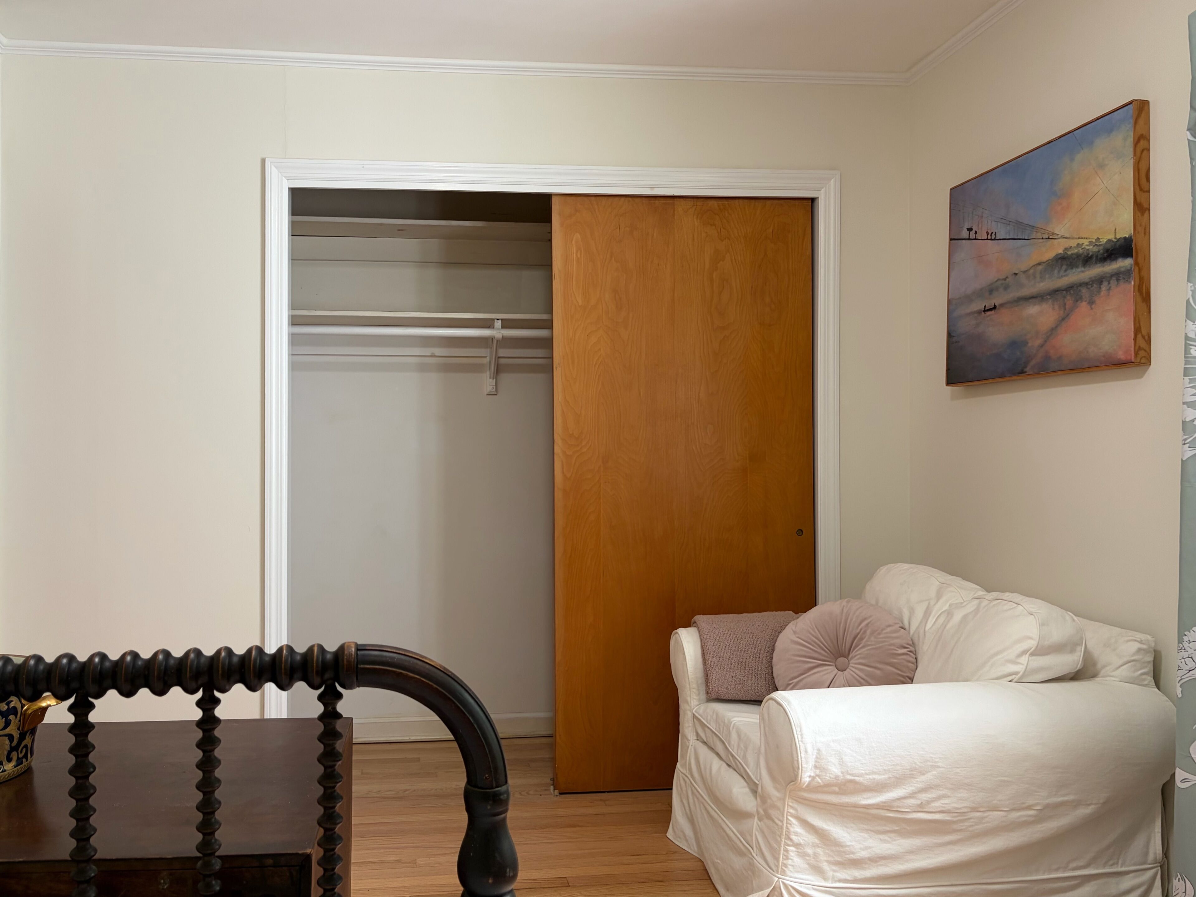 A room with a wooden door, a white sofa, a painting on the wall, and a wooden chair.