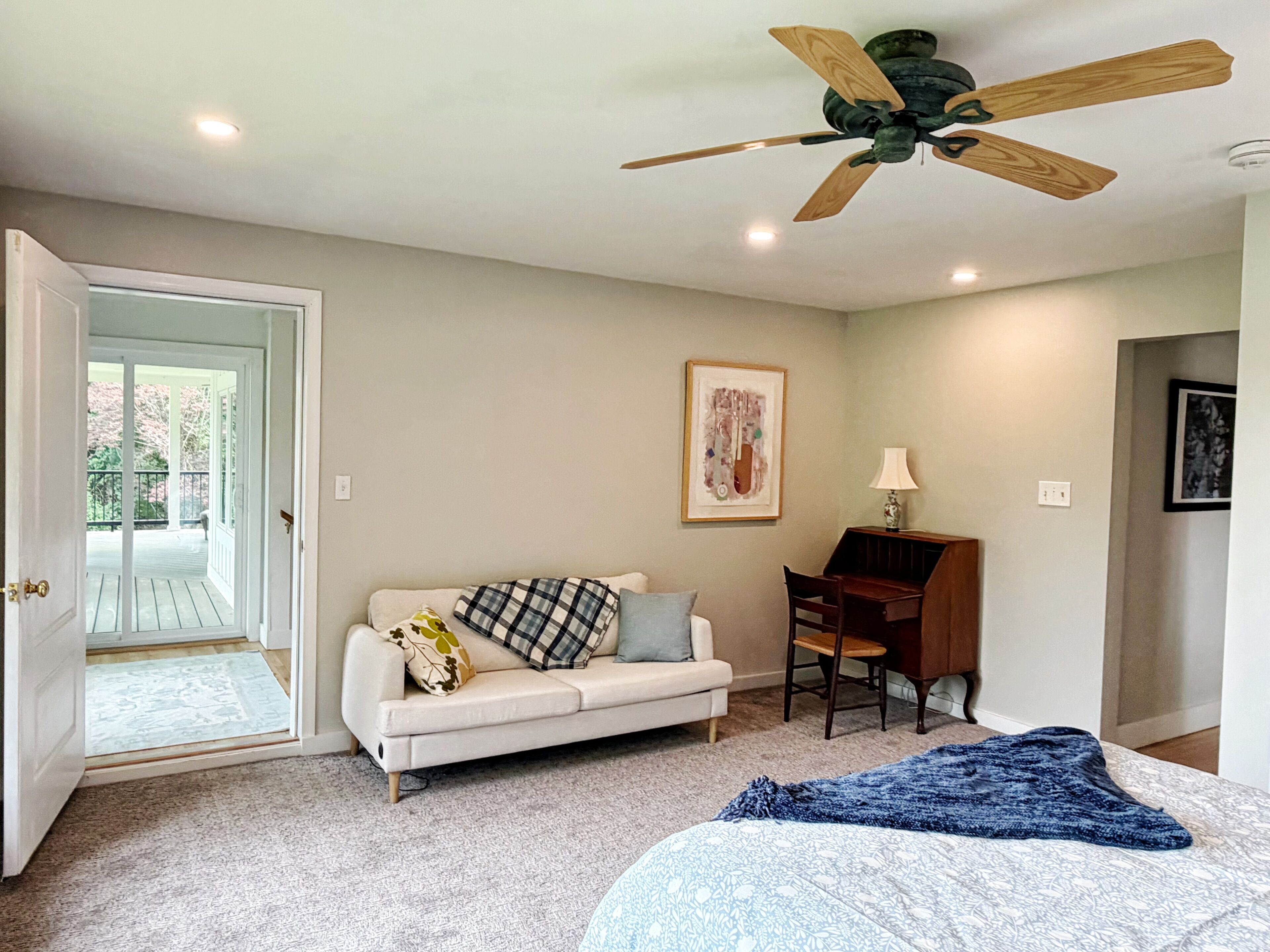 A bedroom with a white sofa, a desk, a ceiling fan, a door leading to a balcony, and a framed picture on the wall.