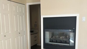 A room with a white door, a fireplace with a glass enclosure, and a kitchen area in the background. - Seaside Cedar Escape (Nanoose Bay)