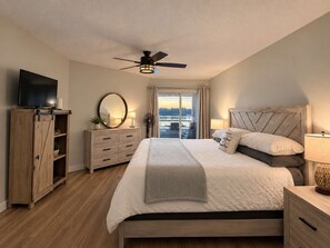 A bedroom with a bed, a television, a ceiling fan, a dresser, and a view of a body of water through a window. - Seaside Cedar Escape (Nanoose Bay)