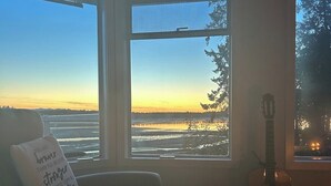 A room with a view of a sunset over a body of water, a guitar, and a chair with a cushion. - Seaside Cedar Escape (Nanoose Bay)