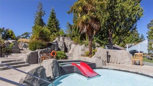 A swimming pool with a red slide, surrounded by rocks and trees. - Seaside Cedar Escape (Nanoose Bay)
