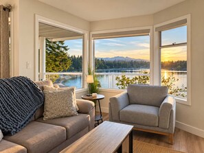 A room with a sofa, armchair, and a view of a lake and mountains. - Seaside Cedar Escape (Nanoose Bay)