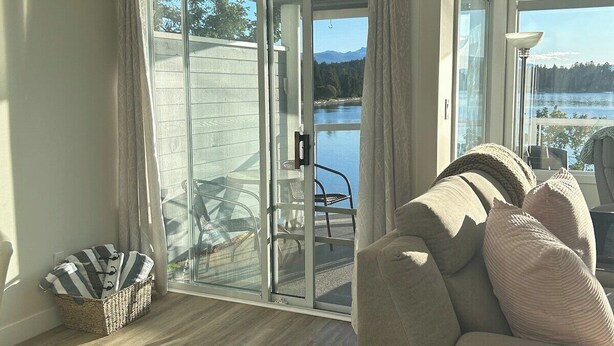 A living room with a sofa, a glass door leading to a balcony, and a view of a lake. - Seaside Cedar Escape (Nanoose Bay)