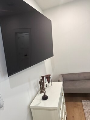 Ein modernes Wohnzimmer mit einem an der Wand befestigten Flachbildfernseher, einer weißen Kommode mit Schubladen, einem grauen Sofa und einem kleinen Tisch mit Dekorationen.