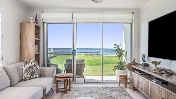 Ein Wohnzimmer mit einer Couch, einem Couchtisch, einem Fernseher und einer Schiebetür, die auf einen Balkon mit Meerblick führt.