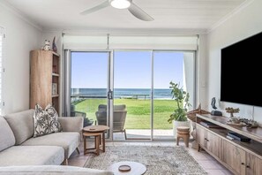 A living room with a sofa, a coffee table, a TV, and a sliding glass door leading to a balcony with a view of the ocean. - Absolute Beachfront Apartment 11B (Lennox Head)