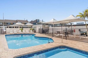 Ein Pool mit Fliesenbordstein, umgeben von Liegestühlen und Sonnenschirmen, mit Gebäuden im Hintergrund.
