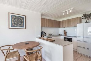 A modern kitchen with a central island, wooden cabinets, and a dining area with a round table and chairs. - Absolute Beachfront Apartment 11B (Lennox Head)