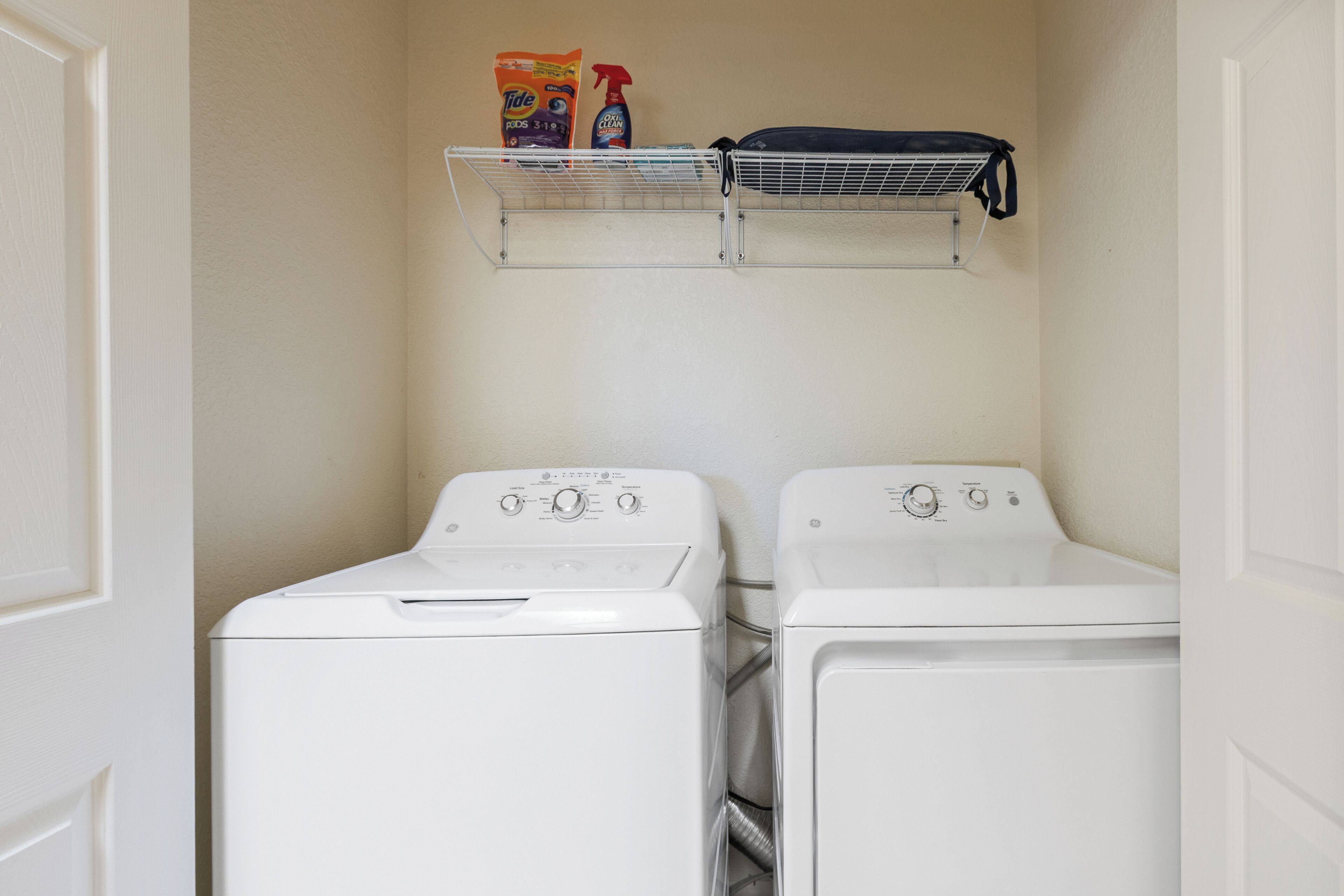 Un cuarto de lavandería con dos lavadoras de carga frontal y un estante superior con productos de limpieza.