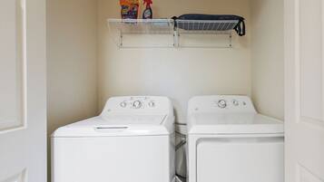 Un cuarto de lavandería con dos lavadoras de carga frontal y un estante superior con productos de limpieza.