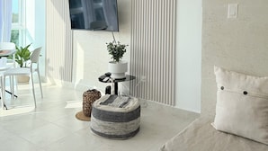 A modern living room with a flat-screen TV mounted on the wall, a standing fan, a potted plant, a round ottoman, and a sofa with a cushion.