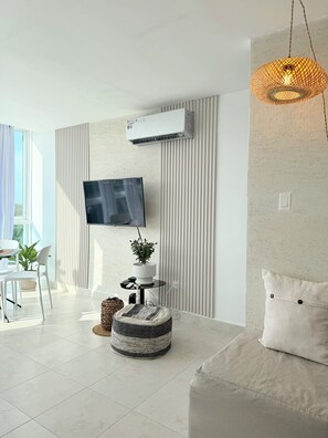 A modern living room with a flat-screen TV mounted on the wall, a standing fan, a potted plant, a round ottoman, and a sofa with a cushion.