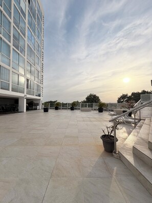 A rooftop terrace with a modern building, potted plants, and outdoor seating.