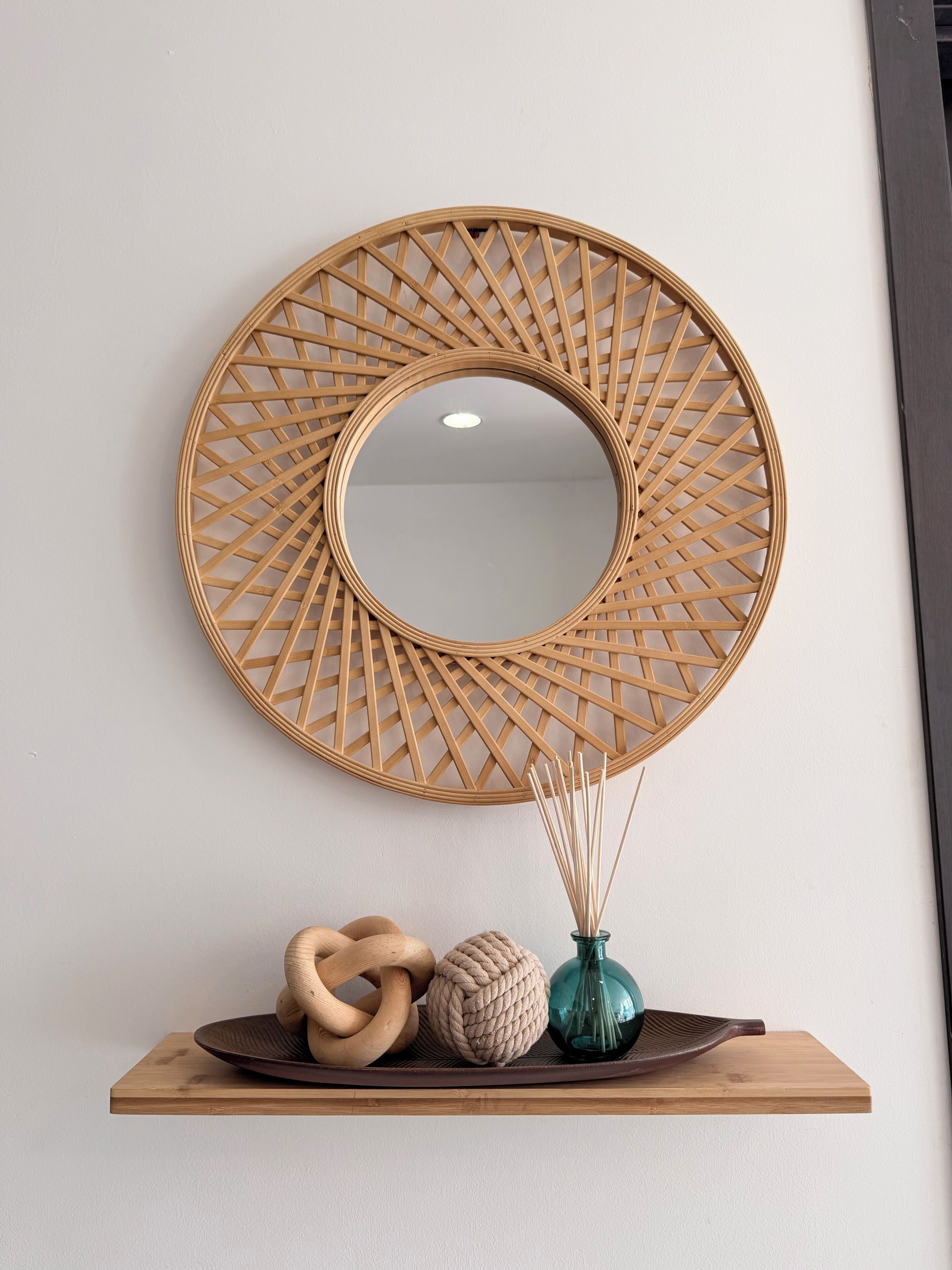 Un miroir rond fixé au mur, doté d’un cadre tressé, une sculpture en bois, une boule texturée et un vase en verre contenant des roseaux, le tout posé sur une étagère en bois.