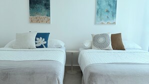 A room with two beds, each with a white bedspread and a grey blanket. There are two wall-mounted paintings, one depicting a beach scene and the other a sky with clouds.