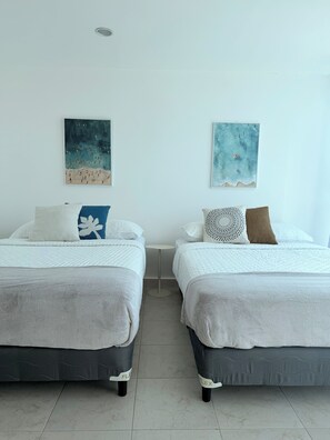A room with two beds, each with a white bedspread and a grey blanket. There are two wall-mounted paintings, one depicting a beach scene and the other a sky with clouds.