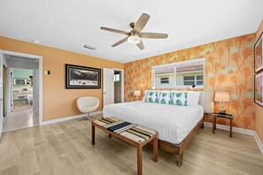 Ein Schlafzimmer mit einem Bett, einem Sessel, einer Bank, einem Deckenventilator und einem Fenster mit Blick auf ein Haus.