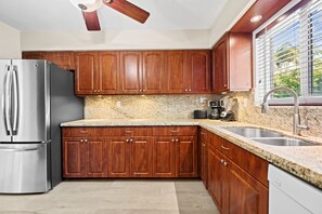 A modern kitchen with wooden cabinets, a stainless steel refrigerator, granite countertops, and a window with blinds. - 1980s Retro Home ~ 12 Minute Walk to Beach: 2BR/2BA with kitchen, WiFi, parking, and patio. (Pompano Beach)