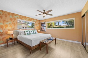 A bedroom with a bed, bedside tables, a ceiling fan, a window with a view, and a wall with a tropical wallpaper pattern. - 1980s Retro Home ~ 12 Minute Walk to Beach: 2BR/2BA with kitchen, WiFi, parking, and patio. (Pompano Beach)