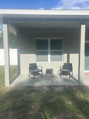 A covered patio area with two chairs and a small table, a window with blinds, and a concrete floor.