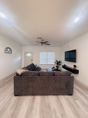 A living room with a dark brown sofa, a ceiling fan, a TV mounted on the wall, and a window with blinds.