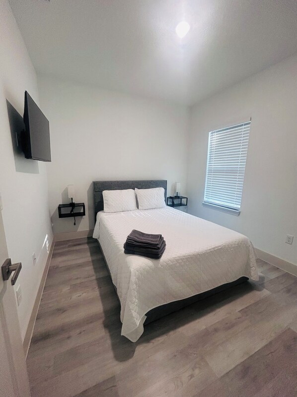 A bedroom with a bed, bedside tables, a TV, and a window with blinds.