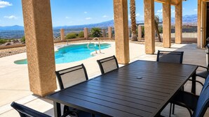 An outdoor pool area with a table and chairs, surrounded by a fence and palm trees, with a view of mountains in the background. - Desert Retreat / Brand new interior / Pool / Views (Bullhead City)